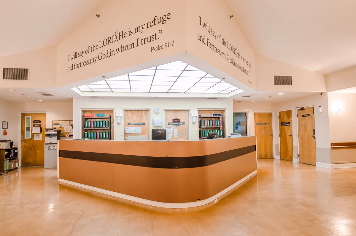 Reception area inside Surrey Place Nursing Center with a large brown desk in the center. Above the desk, a religious quote from Psalm 91:2 is displayed on the wall. Several doors and shelves with files are visible behind the desk. The floor is light wood, and the ceiling has a large light panel.