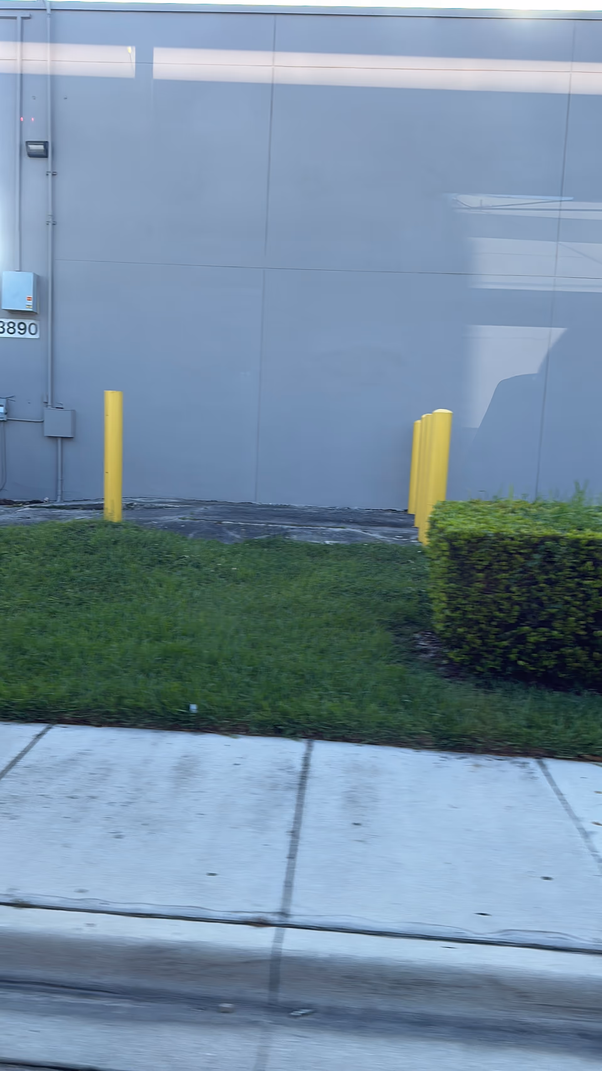 Gray building exterior with yellow bollards, sidewalk, grass, and a trimmed hedge.