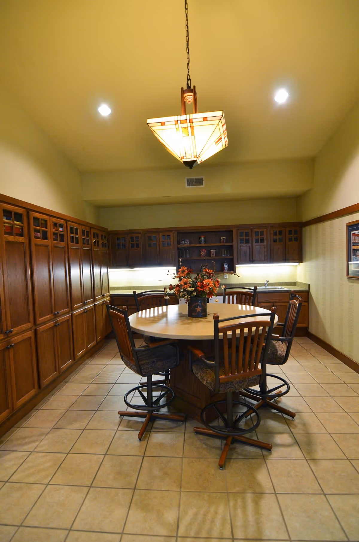 A kitchen area with wooden cabinets lining the walls, a round table in the center surrounded by five cushioned chairs with wooden backs, and a decorative flower arrangement on the table. The room has tiled flooring and a hanging light fixture above the table.