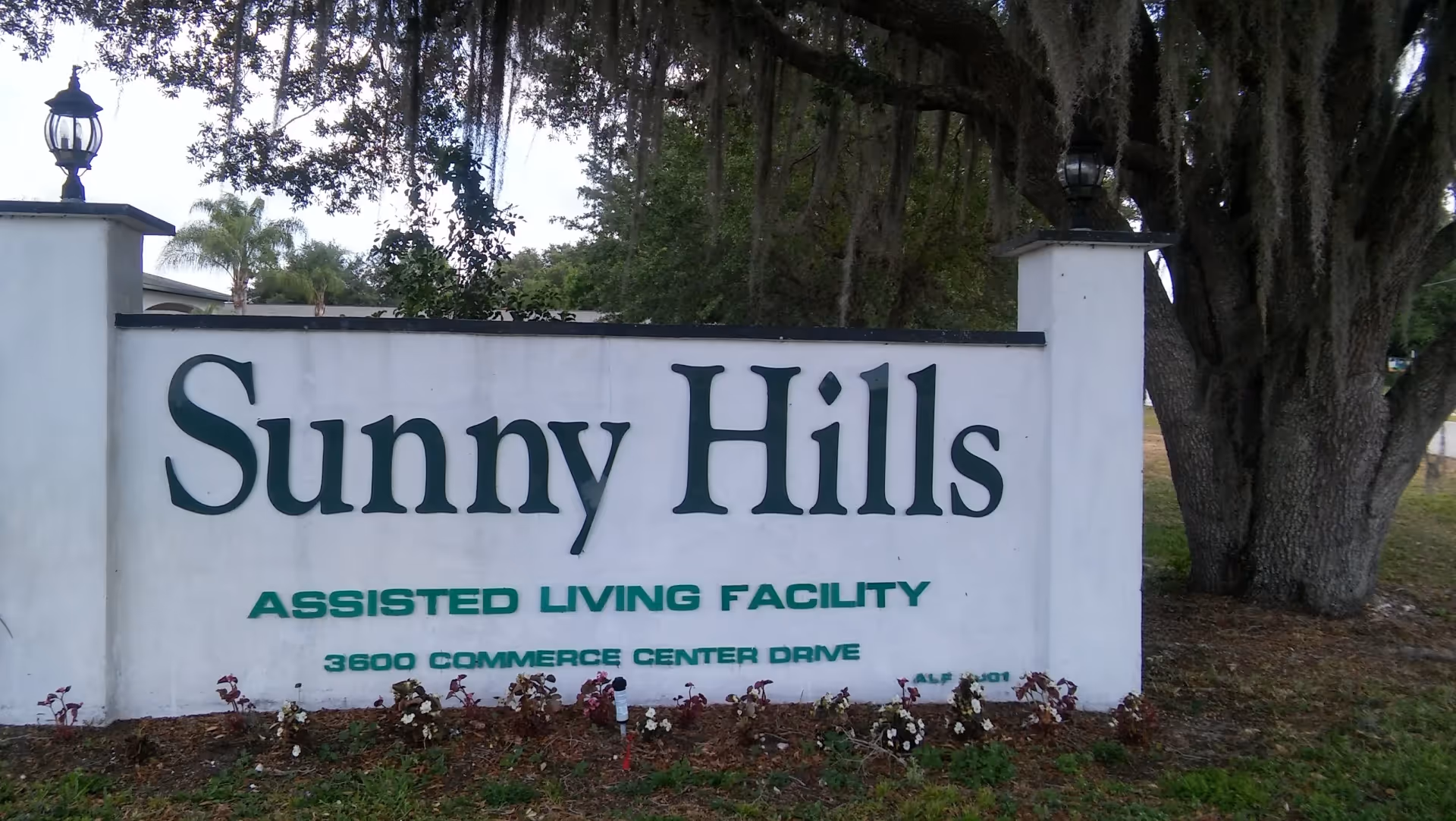 A large white sign with green text reading 'Sunny Hills Assisted Living Facility 3600 Commerce Center Drive' situated outdoors with trees and plants around it.