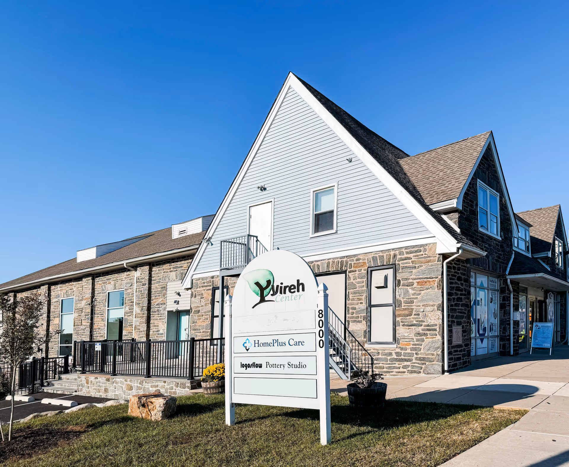 Stone-and-siding senior living building with a curved sign, stairs, and sidewalk under a clear blue sky.