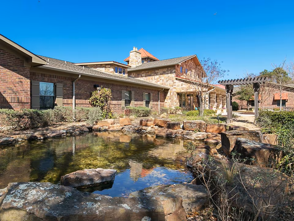 Stone-and-brick assisted living building with a landscaped pond and rock garden in the foreground under a clear blue sky.