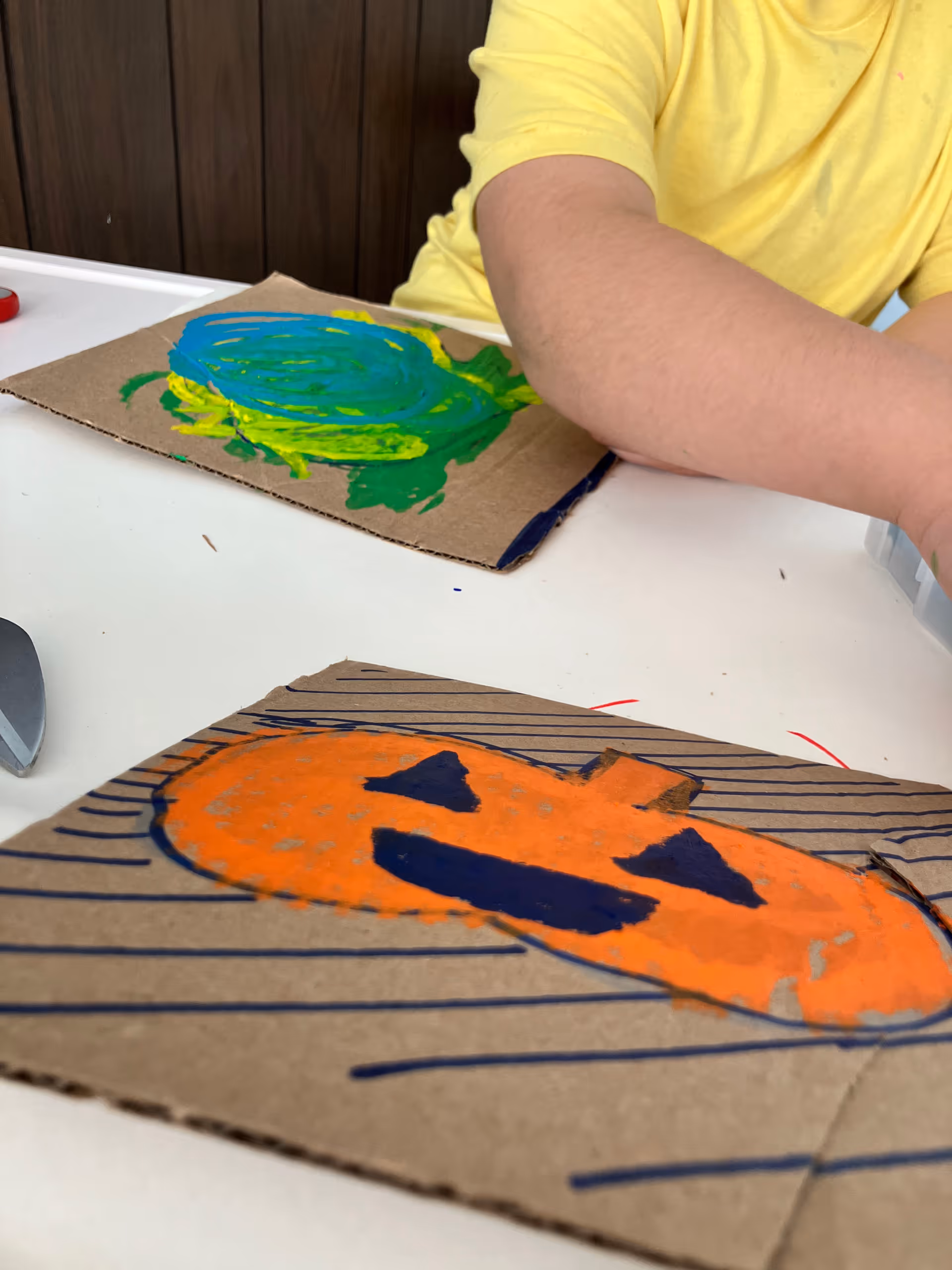 Person in a yellow shirt paints orange and blue shapes on cardboard at a table indoors.