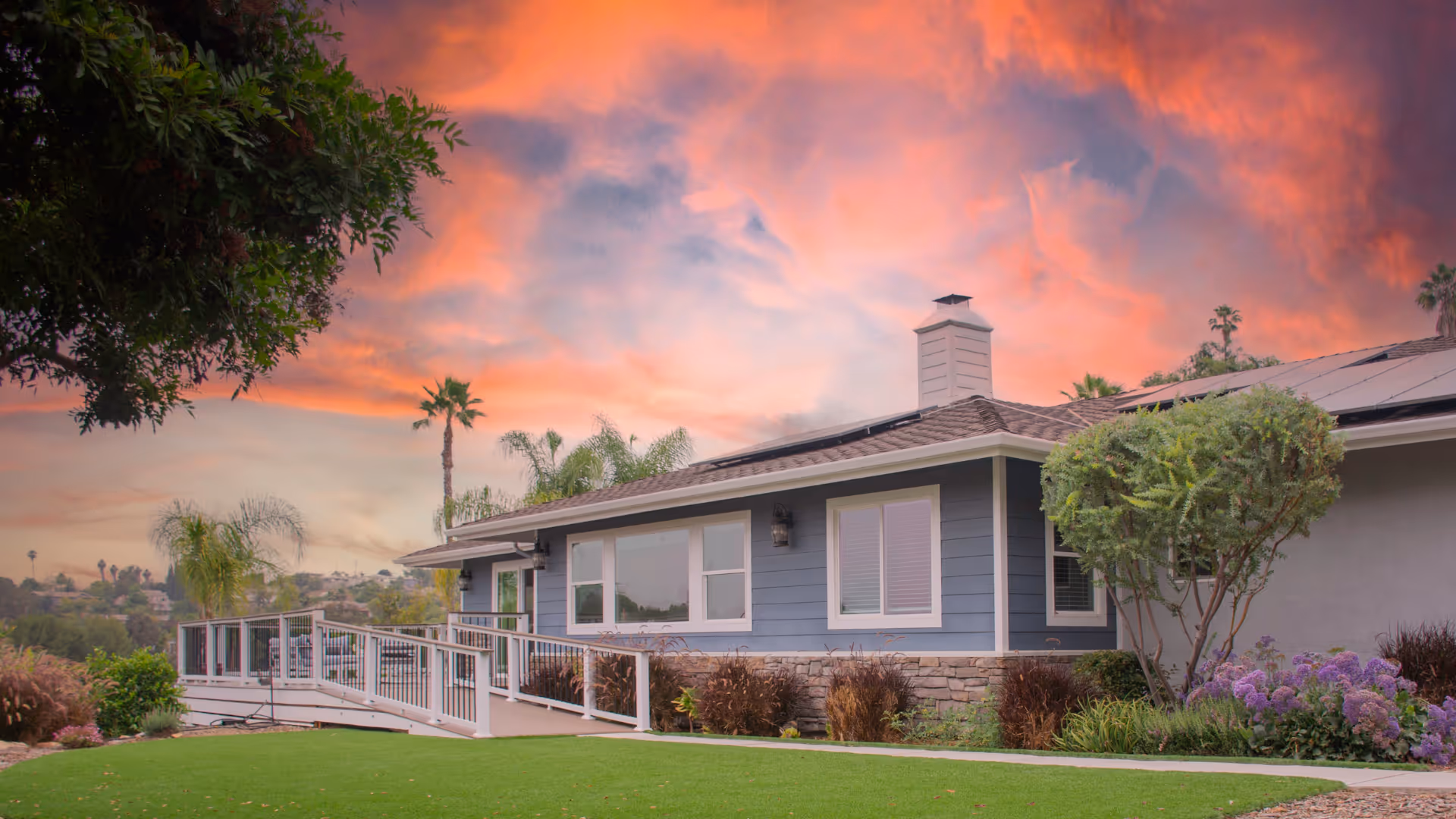 Single-story blue assisted living building with a ramp and sunset sky.