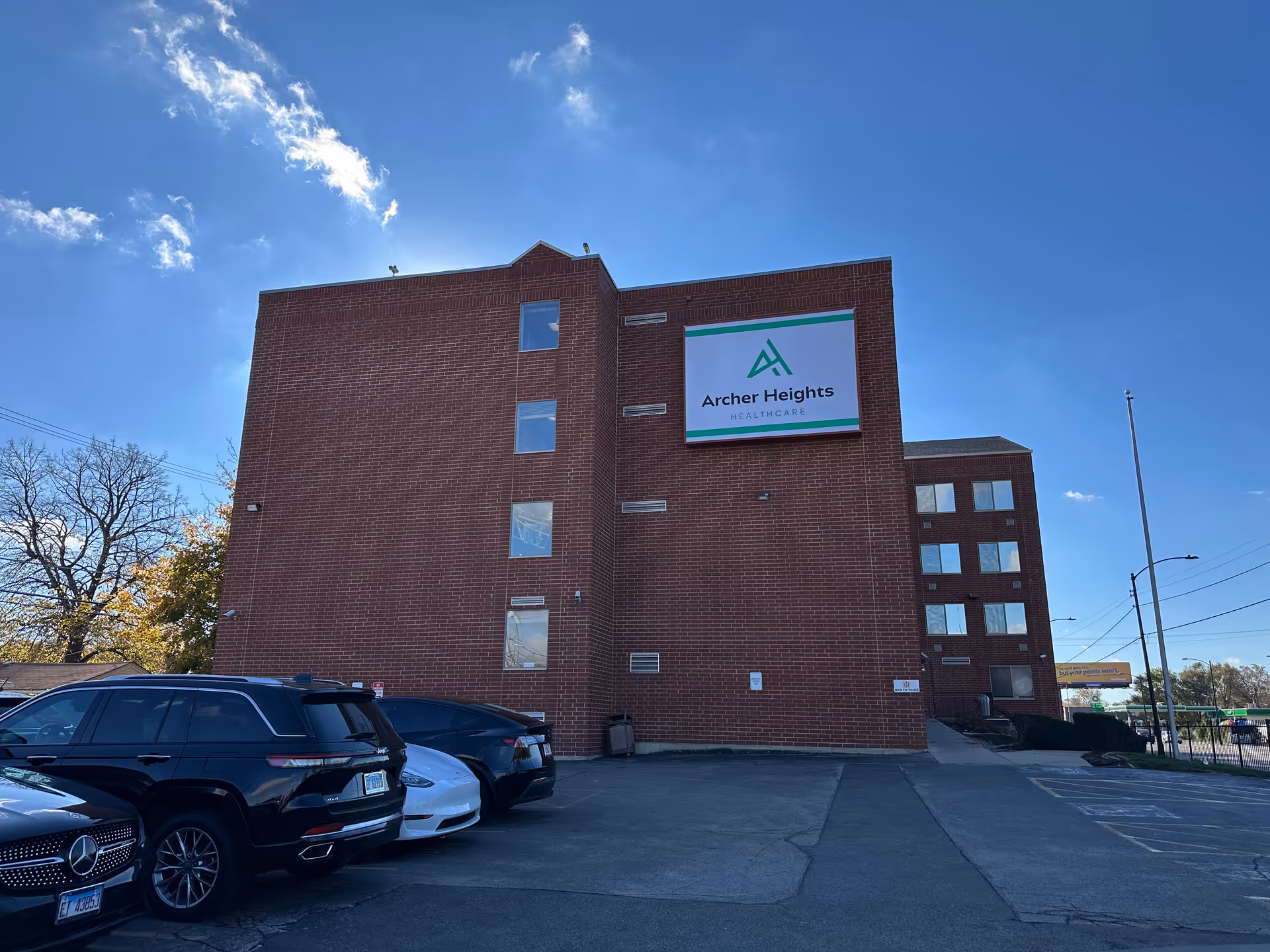 Brick multi-story healthcare building with an Archer Heights Healthcare sign and parked cars in front under a blue sky.