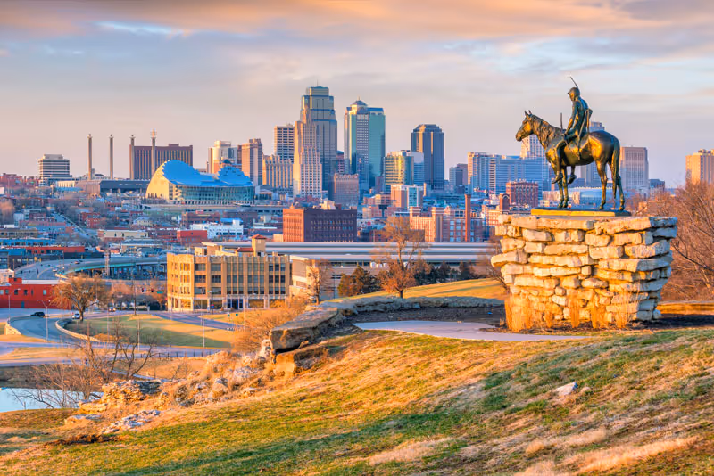 View of the Kansas City skyline at sunset with a bronze statue of a mounted rider on a stone pedestal in the foreground, overlooking a grassy hill and urban buildings.