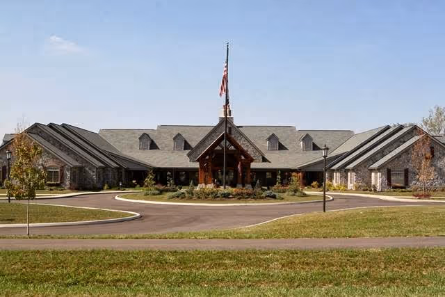 Front exterior view of The Inn at Whitewood Village, a single-story building with a peaked roof and multiple dormer windows. The entrance features a wooden portico with an American flag flying on a flagpole in front. The building is surrounded by a circular driveway and landscaped greenery under a clear blue sky.