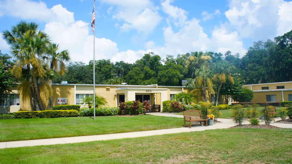 Single-story yellow senior care building with an American flagpole, benches, palm trees, and a landscaped lawn under a blue sky.