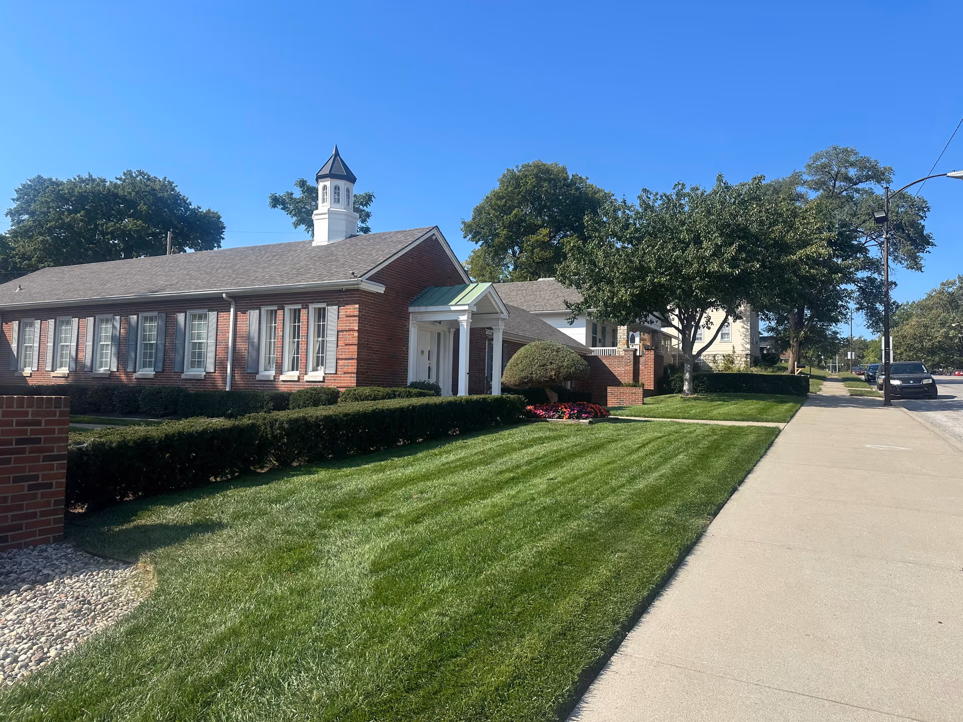 Exterior of a brick senior living facility with manicured lawn and trees along a sidewalk