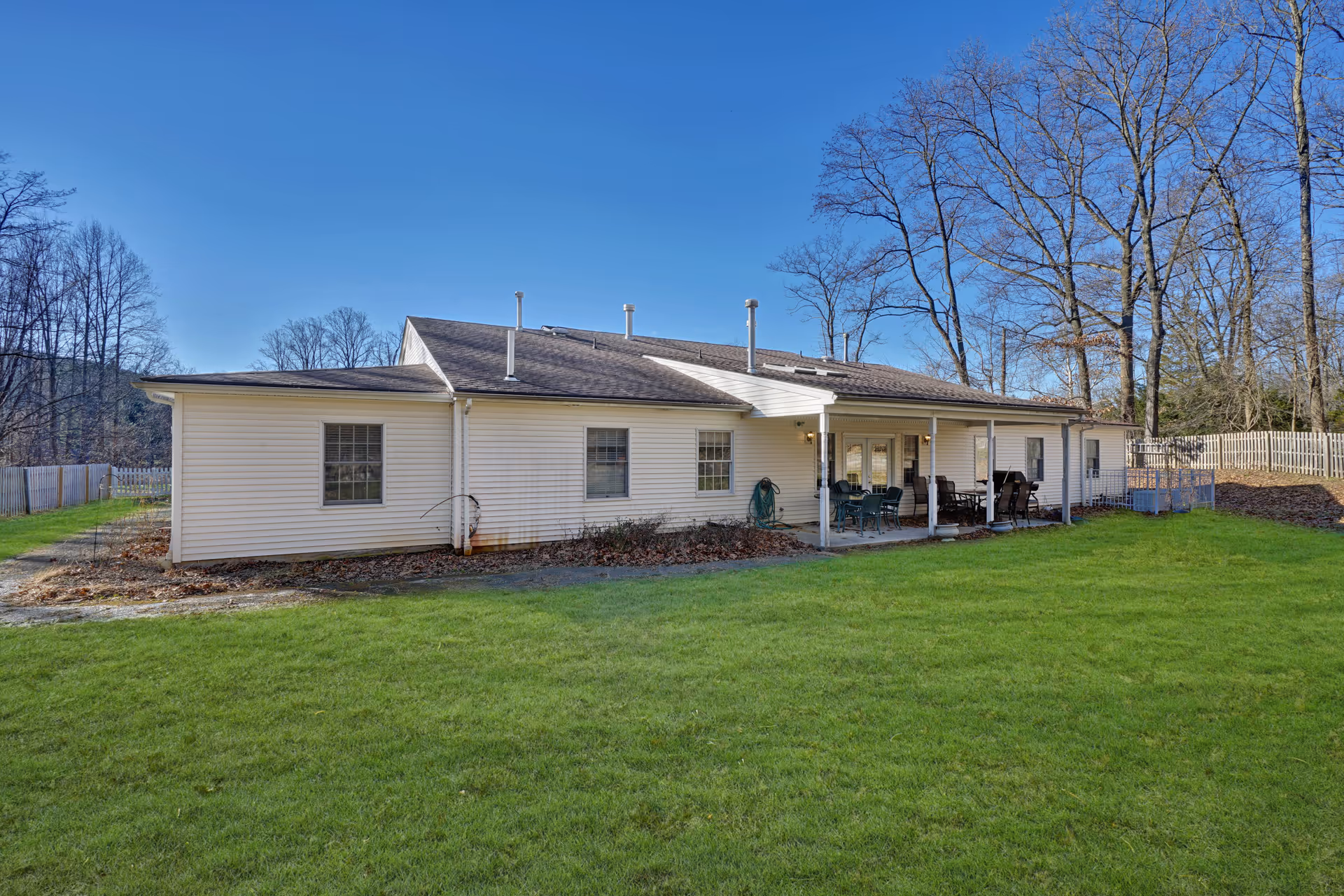 Exterior view of a single-story white building with a covered patio area furnished with outdoor chairs and tables, surrounded by a grassy lawn and leafless trees under a clear blue sky.
