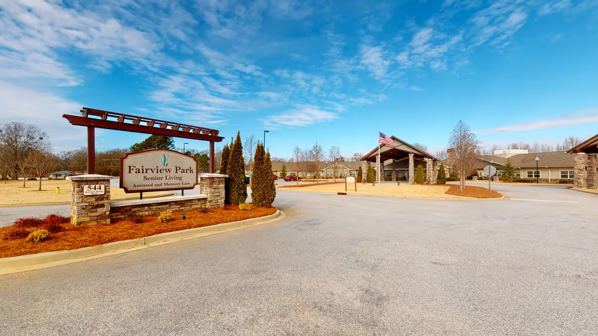 Exterior view of Fairview Park Senior Living facility showing the entrance sign, driveway, and building with an American flag on a clear day.