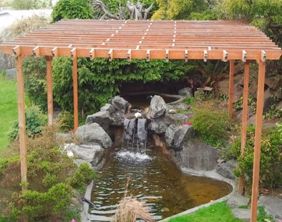 Wooden pergola over a small pond with rocks and lush garden.