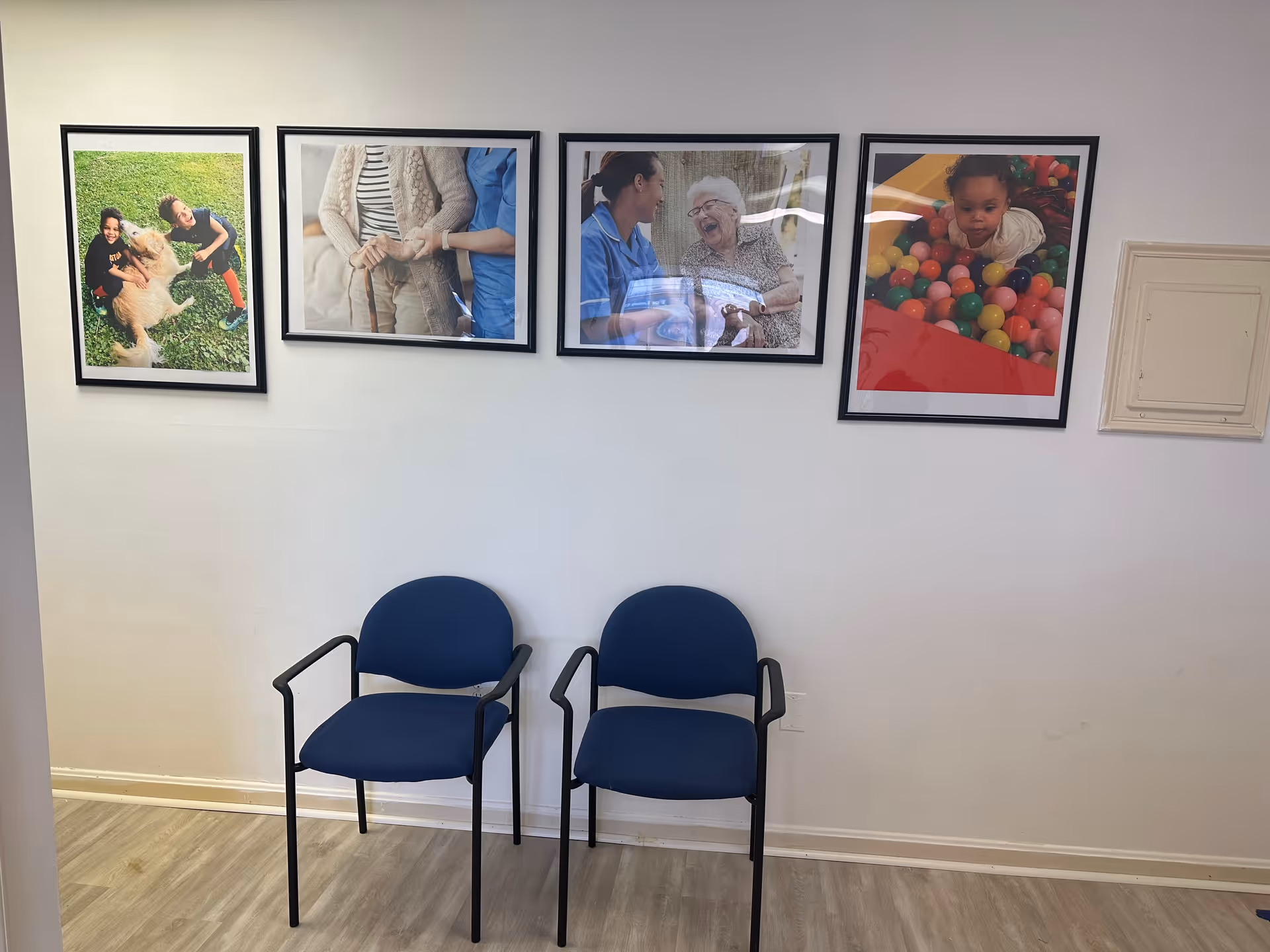 Two blue chairs sit along a wall with four framed photos in a senior living hallway.