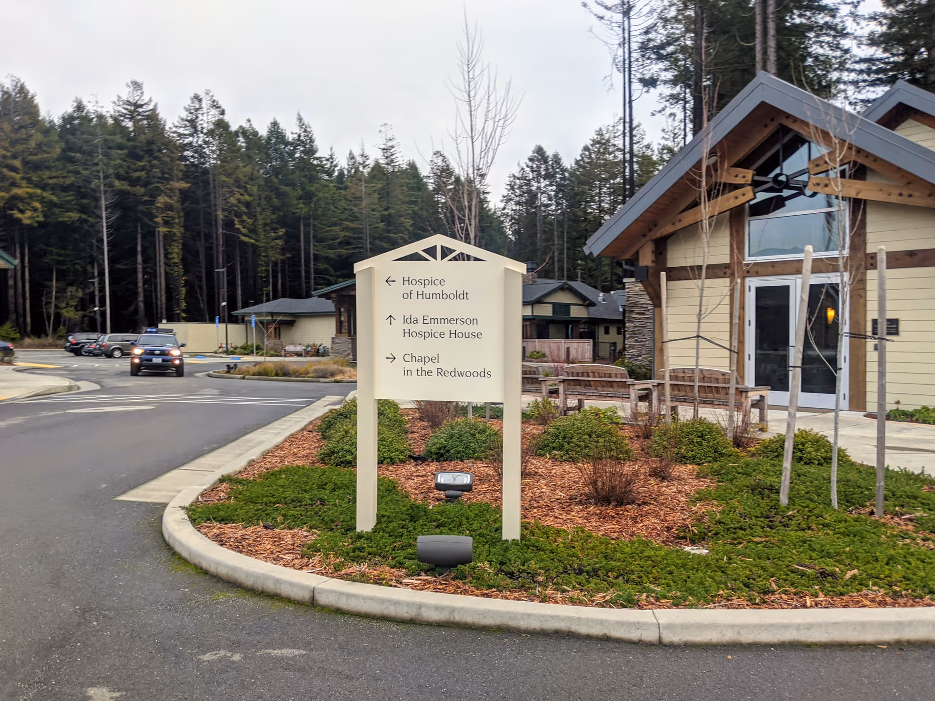 Exterior view of Hospice of Humboldt campus with sign, benches, and wooded backdrop.
