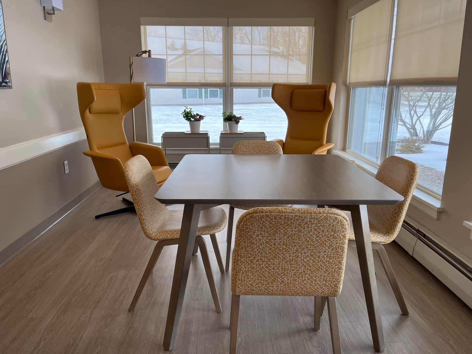 Bright seating area with a square table surrounded by four patterned chairs and two mustard wingback armchairs near large windows.