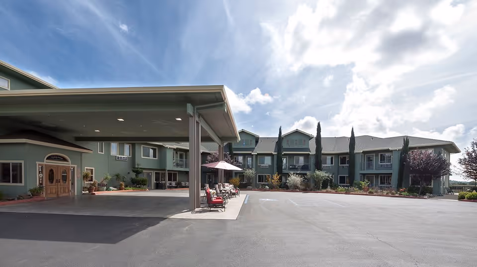 Exterior view of Foothill Village Senior Living facility showing a two-story green building with multiple windows and balconies, a covered entrance with seating and umbrellas, surrounded by landscaped plants and trees under a partly cloudy sky.