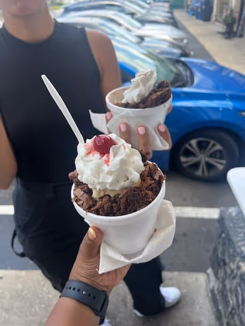 Two people hold ice cream sundaes outdoors near parked cars.