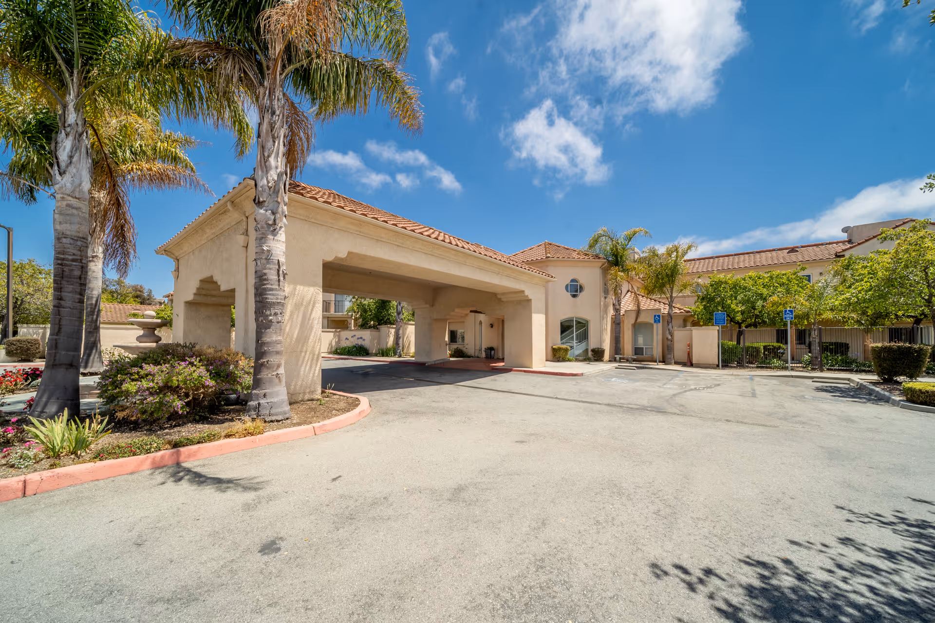 Beige senior living facility exterior with a covered entrance, palm trees, and blue sky.