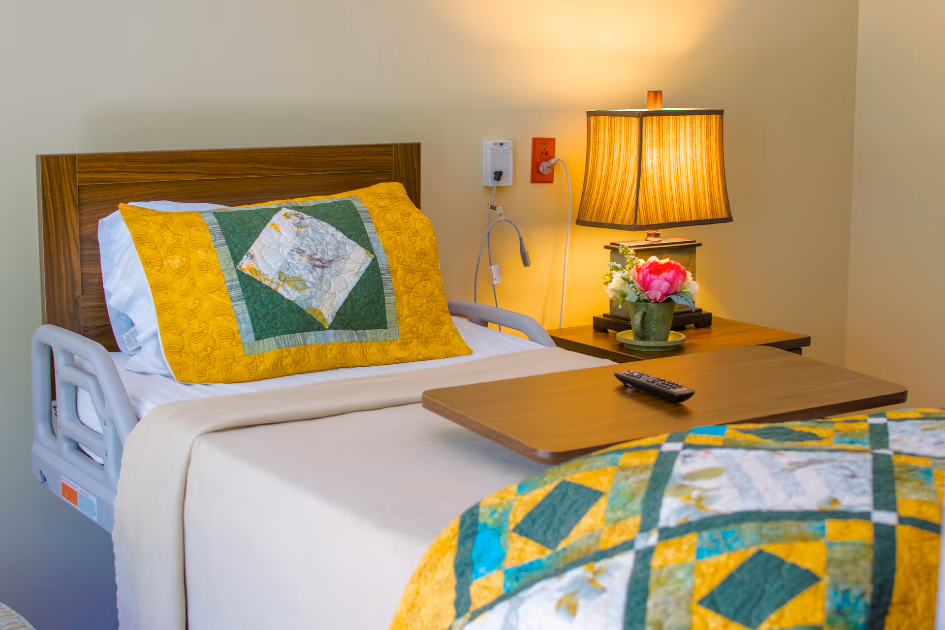 Hospice bedroom with a bed, quilt, and a bedside table with lamp.
