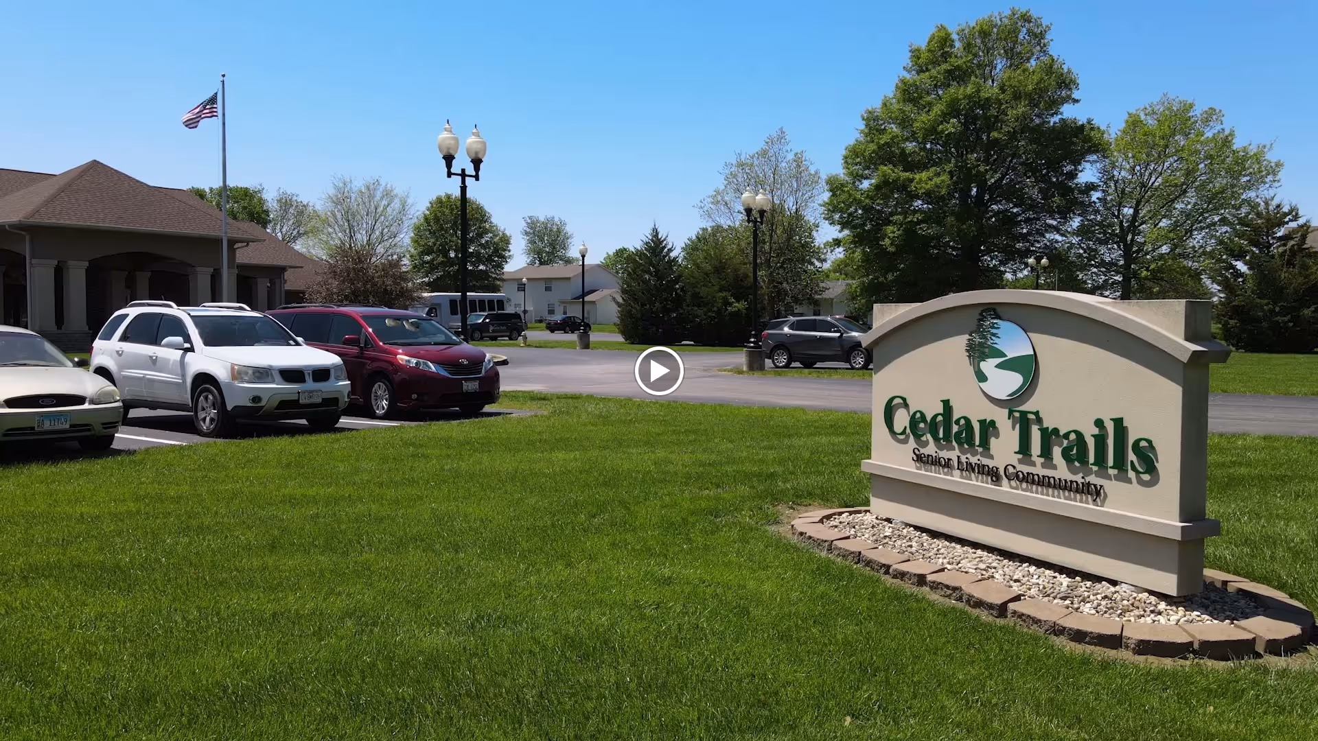 Outdoor view of Cedar Trails Senior Living community sign on a grassy area with a parking lot and several parked cars in the background, along with trees and a building under a clear blue sky.