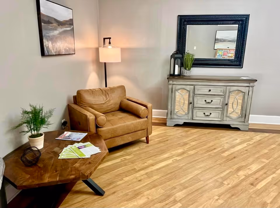 A cozy seating area with a brown leather armchair, wooden side table and lamp, a decorative console with mirror, and a potted plant on hardwood floors.