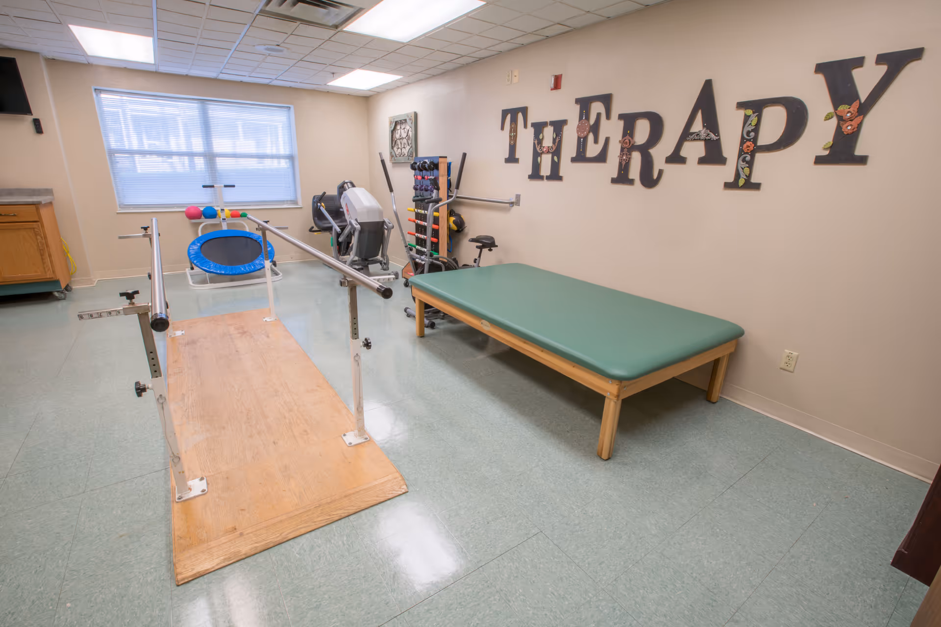 Therapy room with parallel bars, a green treatment table, exercise equipment and large "THERAPY" letters on the wall.