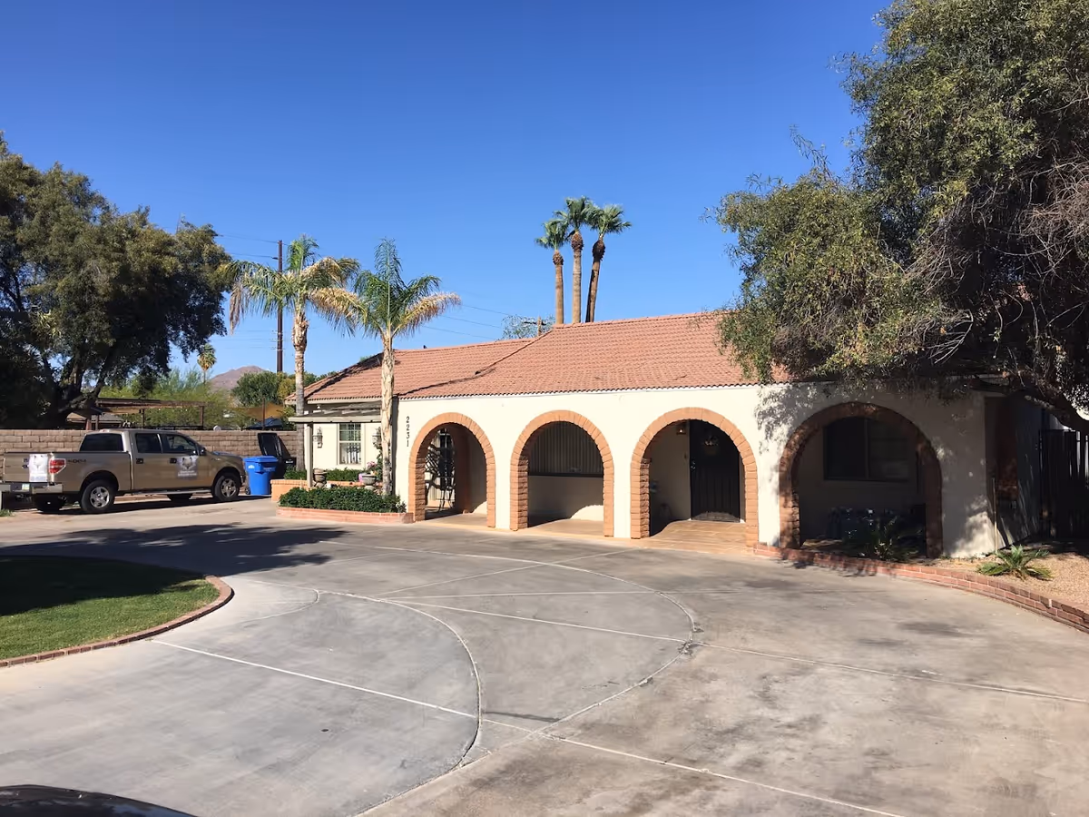 Exterior view of a single-story building with a tiled roof and three arched entryways. There are palm trees and other greenery around the building, a paved driveway in front, and two pickup trucks parked to the left side. The sky is clear and blue.