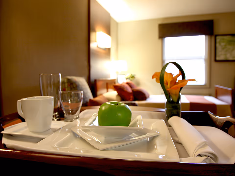 A close-up view of a tray with a green apple on a white plate, a white coffee cup, empty glasses, a rolled white napkin, and a small vase with orange flowers in a softly lit bedroom with a bed and pillows in the background.