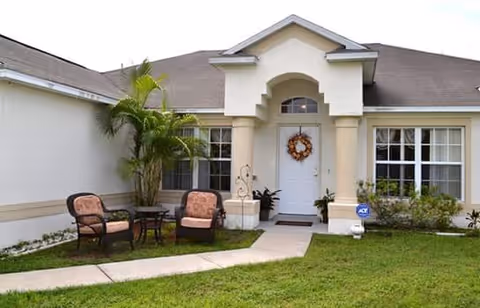 Front entrance of a single-story assisted living home with a wreath on the door, two patio chairs and a small lawn.