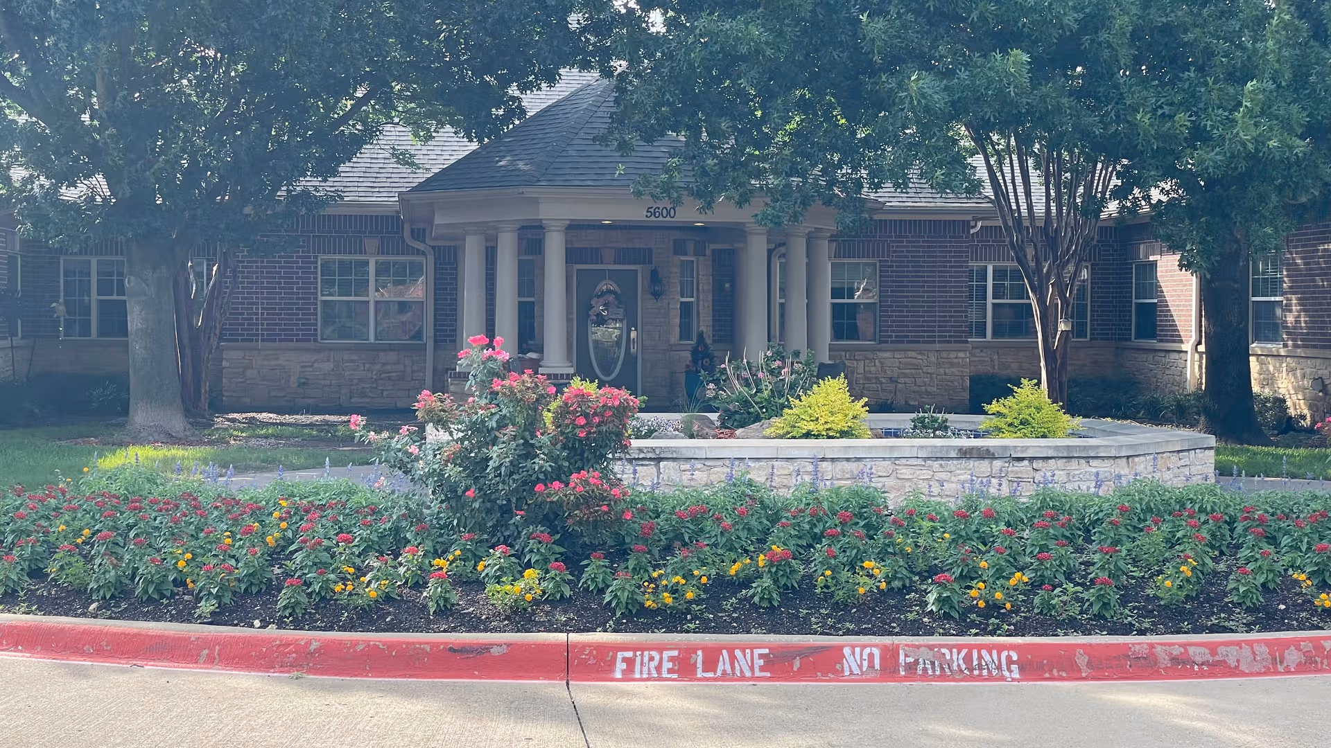 Brick senior living facility entrance with colorful flower beds and a red fire lane curb.