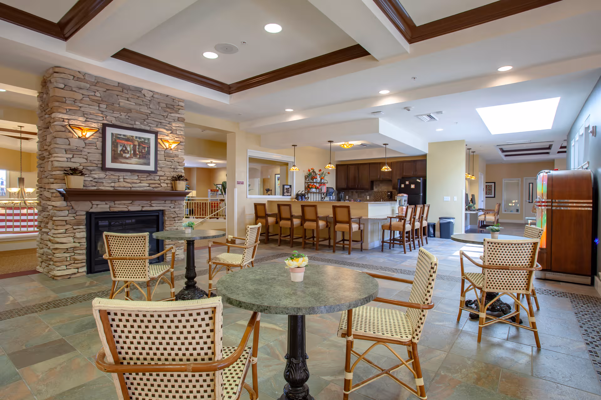 Spacious senior living lounge with stone fireplace, round tables, and a bar counter kitchen area.