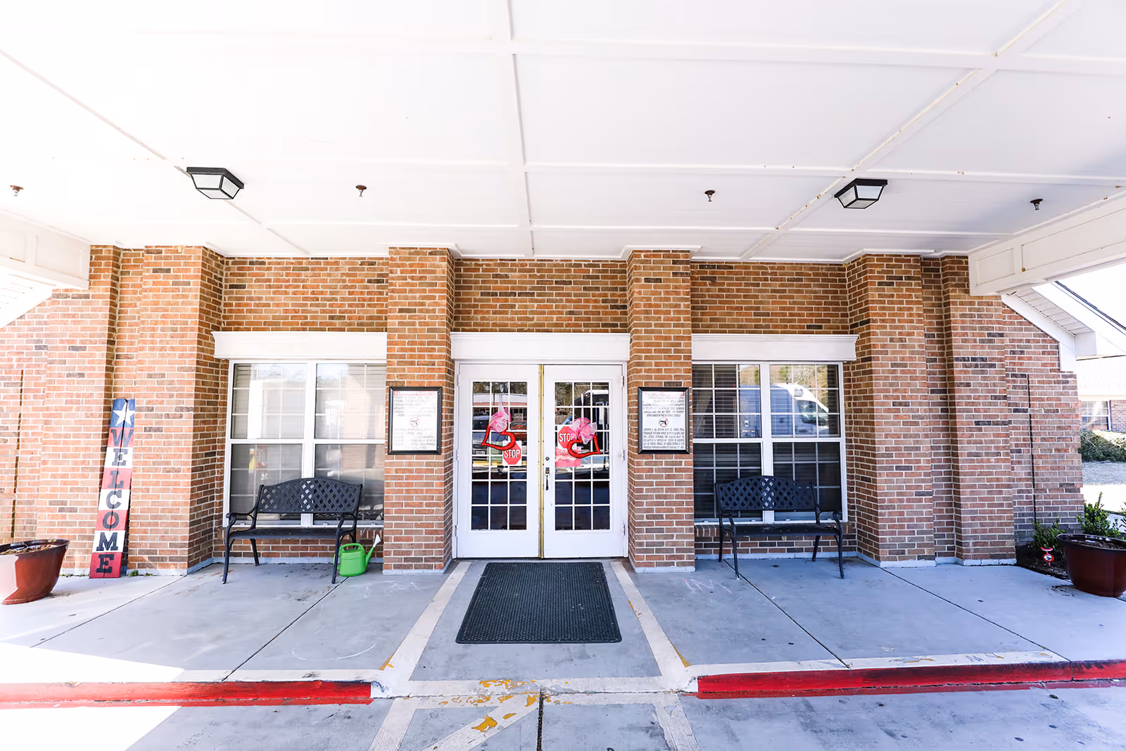 Entrance of Village Creek Rehabilitation & Nursing Center showing double glass doors with stop signs, flanked by two black metal benches on a concrete porch under a white ceiling. The building exterior is made of red brick with white trim. There are potted plants on either side and a vertical wooden welcome sign on the left.