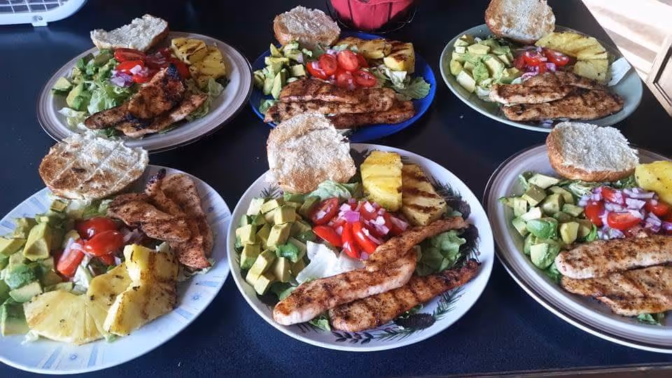Six plates with grilled chicken, salad, avocado, grilled pineapple, and open sandwich buns arranged on a countertop.