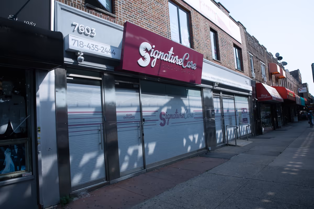 Wide street view of a Signature Care storefront with closed metal shutters along the sidewalk.