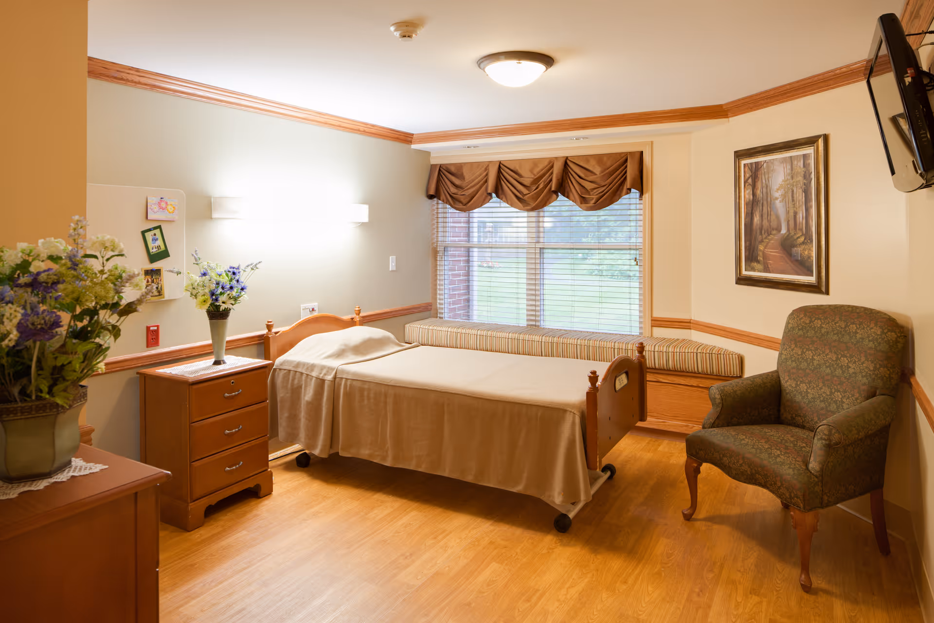 A furnished senior living bedroom with a single bed, nightstand, armchair, window seat, and floral decorations.