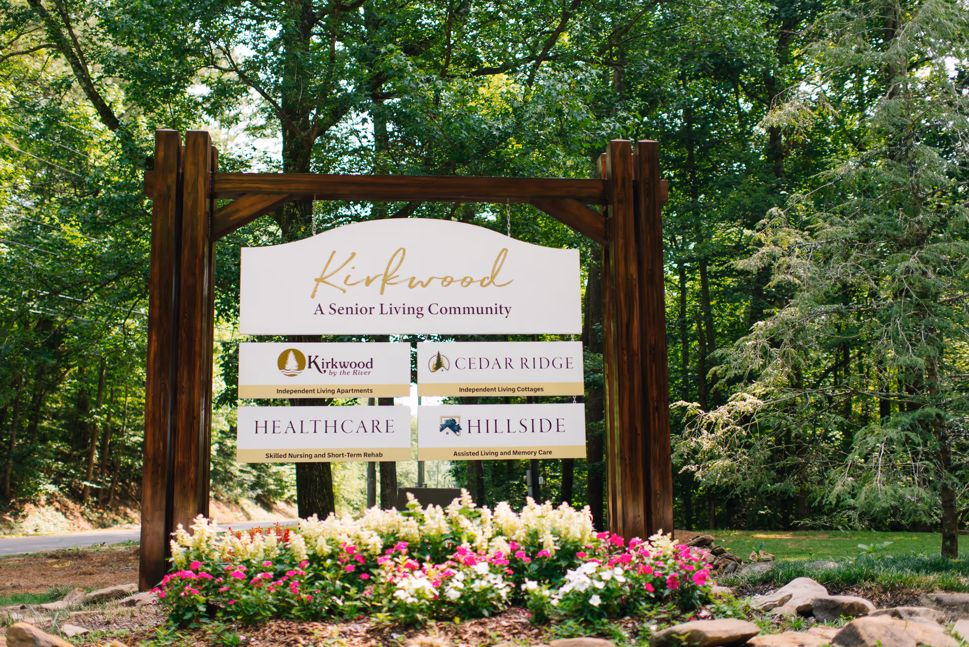 A large wooden signpost in a landscaped garden area with colorful flowers in front. The sign reads 'Kirkwood A Senior Living Community' with additional smaller signs below indicating different living options: Kirkwood by the River Independent Living Apartments, Cedar Ridge Independent Living Cottages, Healthcare Skilled Nursing and Short-Term Rehab, and Hillside Assisted Living and Memory Care. The background shows a wooded area with tall trees.