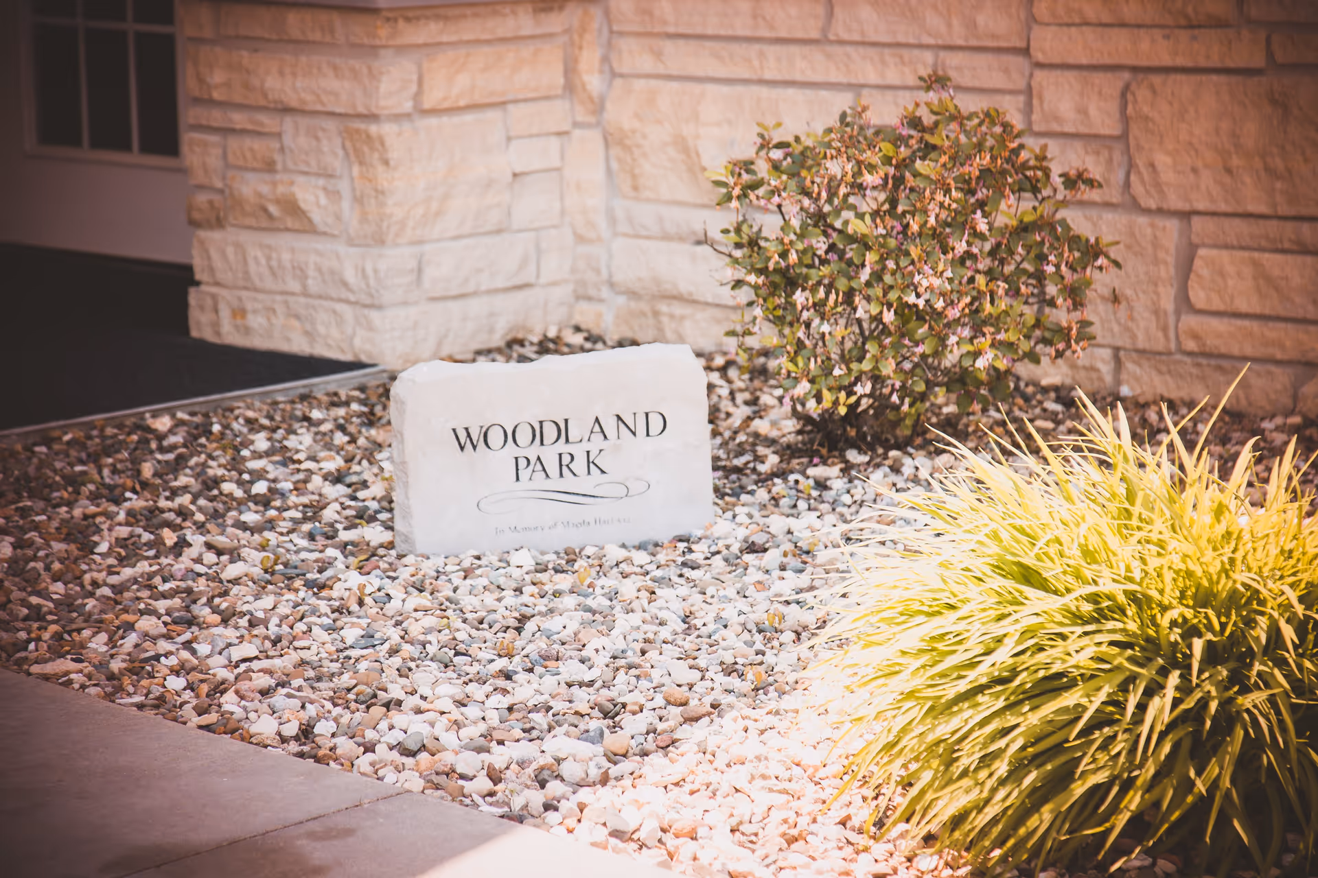 A stone marker engraved with 'WOODLAND PARK' placed in a bed of small rocks and surrounded by green shrubs, next to a stone wall and a doorway.