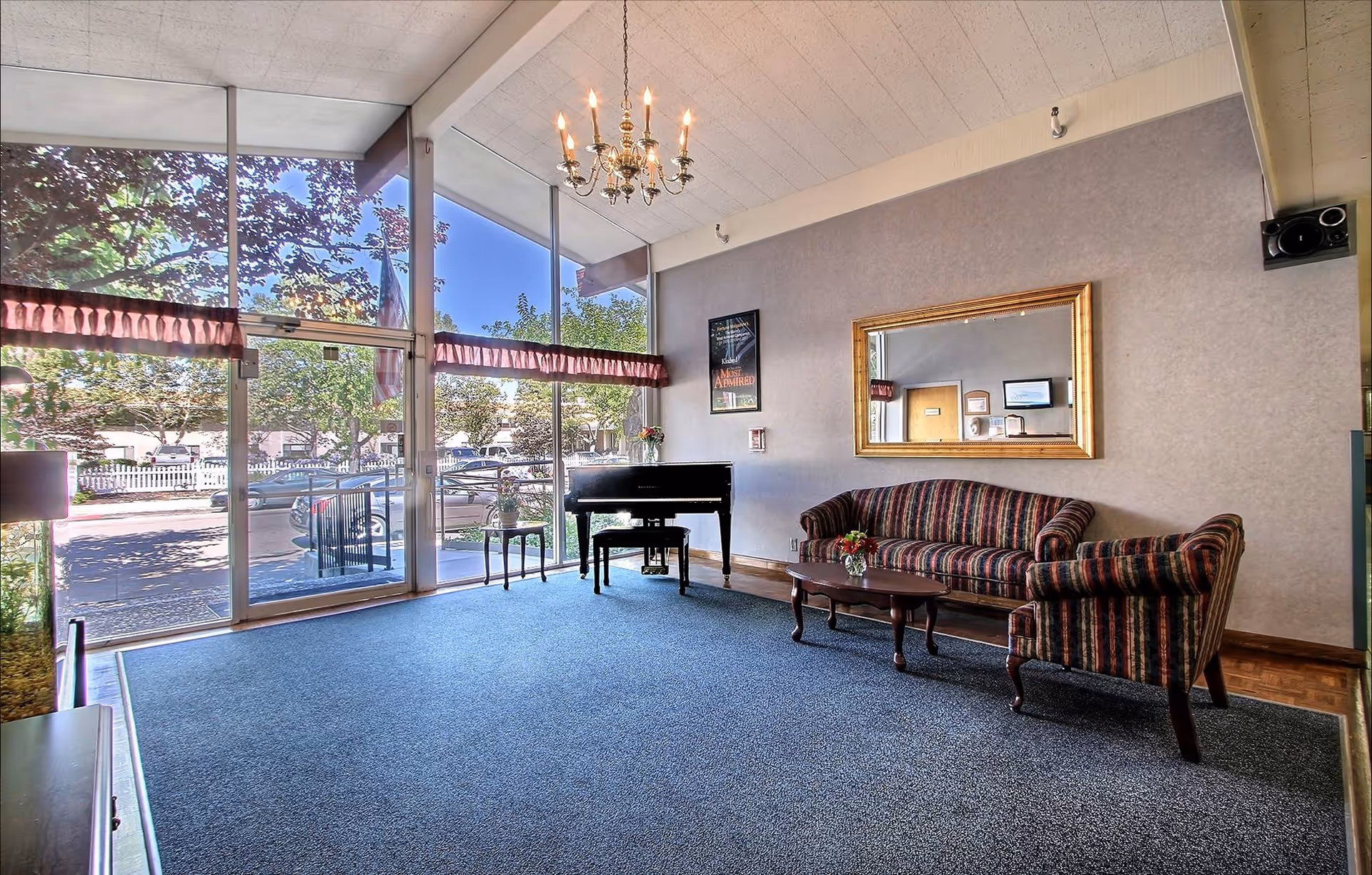 Sunlit senior living lounge with large windows, piano, sofa set, and a chandelier.