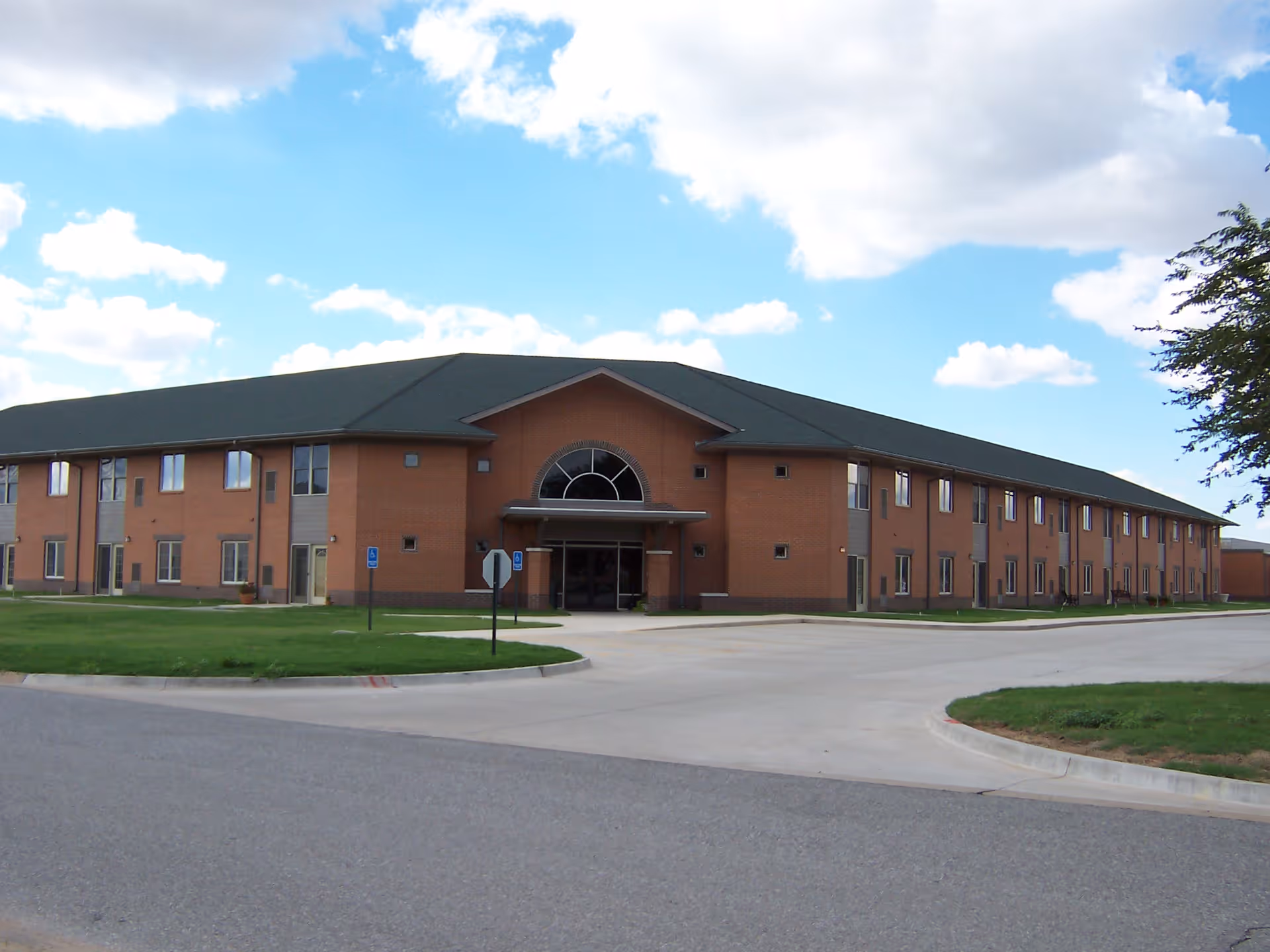Exterior view of a two-story brick building with a dark roof under a partly cloudy blue sky. The building has multiple windows and a main entrance with a covered awning. There is a paved driveway and green lawn surrounding the building.