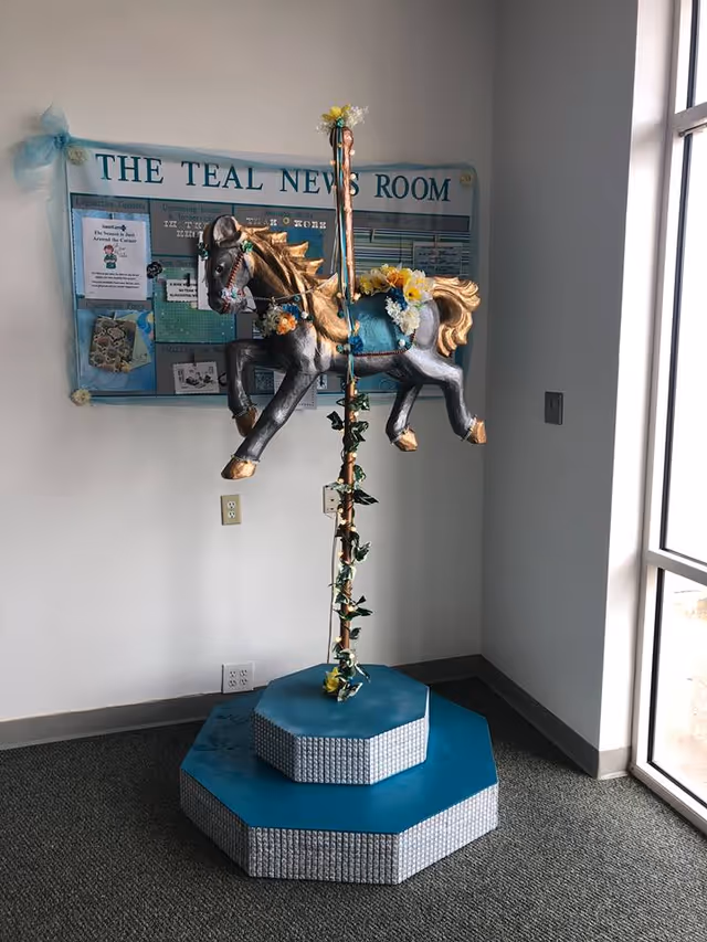 Decorative carousel horse on a tall pedestal in a hallway near a bulletin board.
