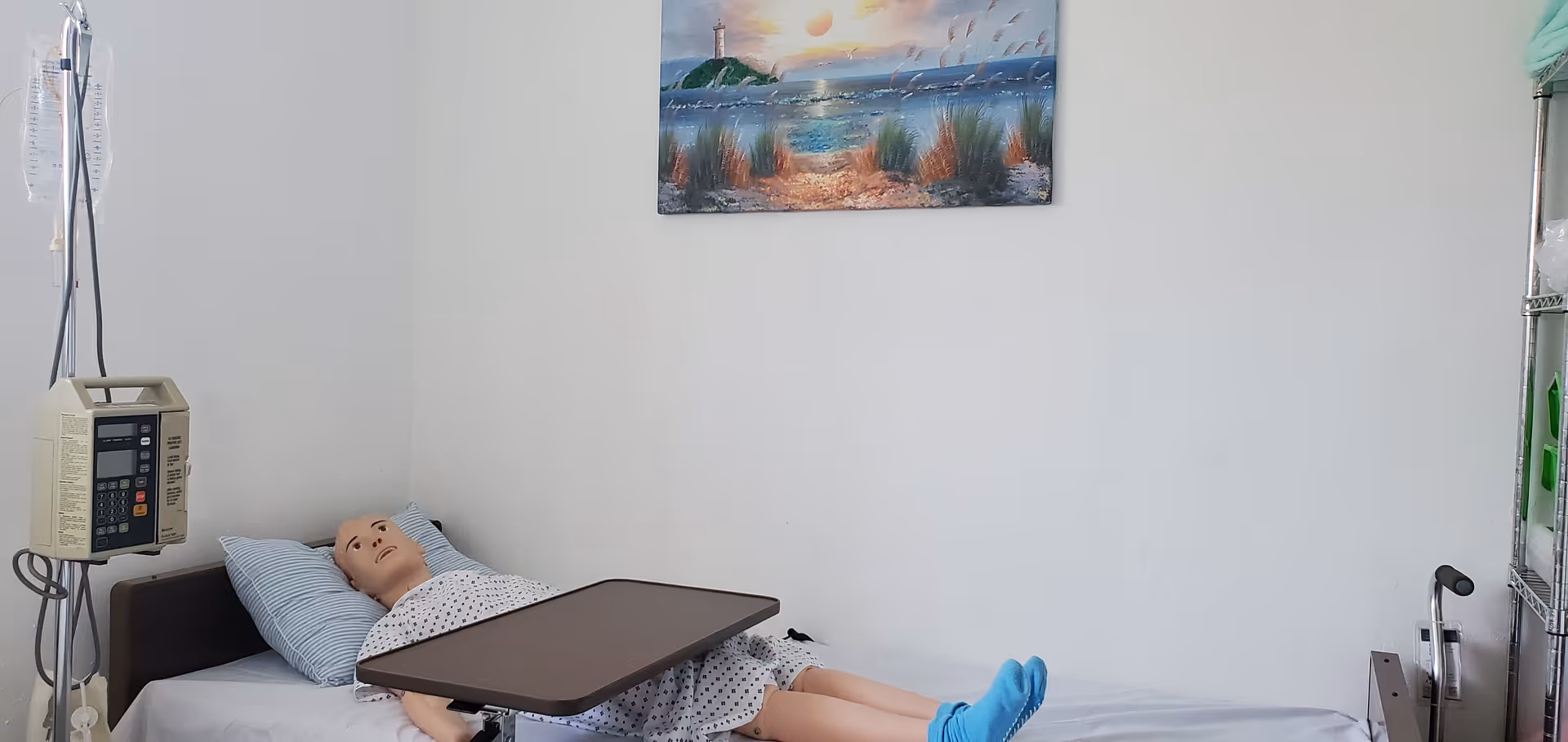 Hospital-style patient bed with a mannequin, IV pump, and tray table in a clean room.