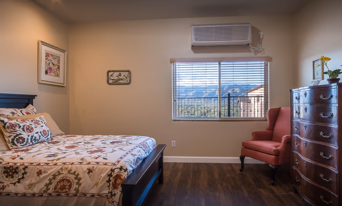 Furnished bedroom with a bed, dresser, red chair, and window view.