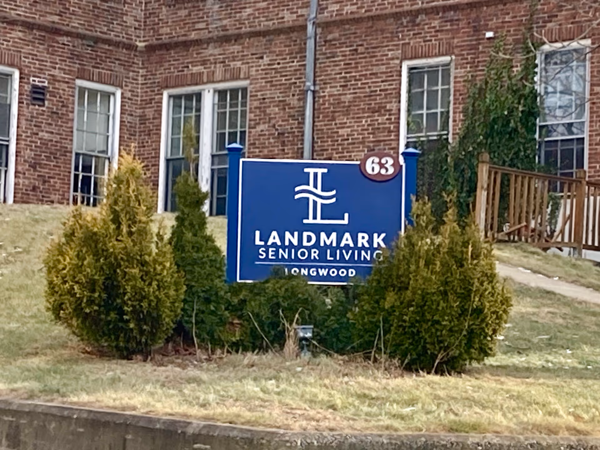 Blue Landmark Senior Living sign on a grassy lawn in front of a brick building with shrubs and windows.
