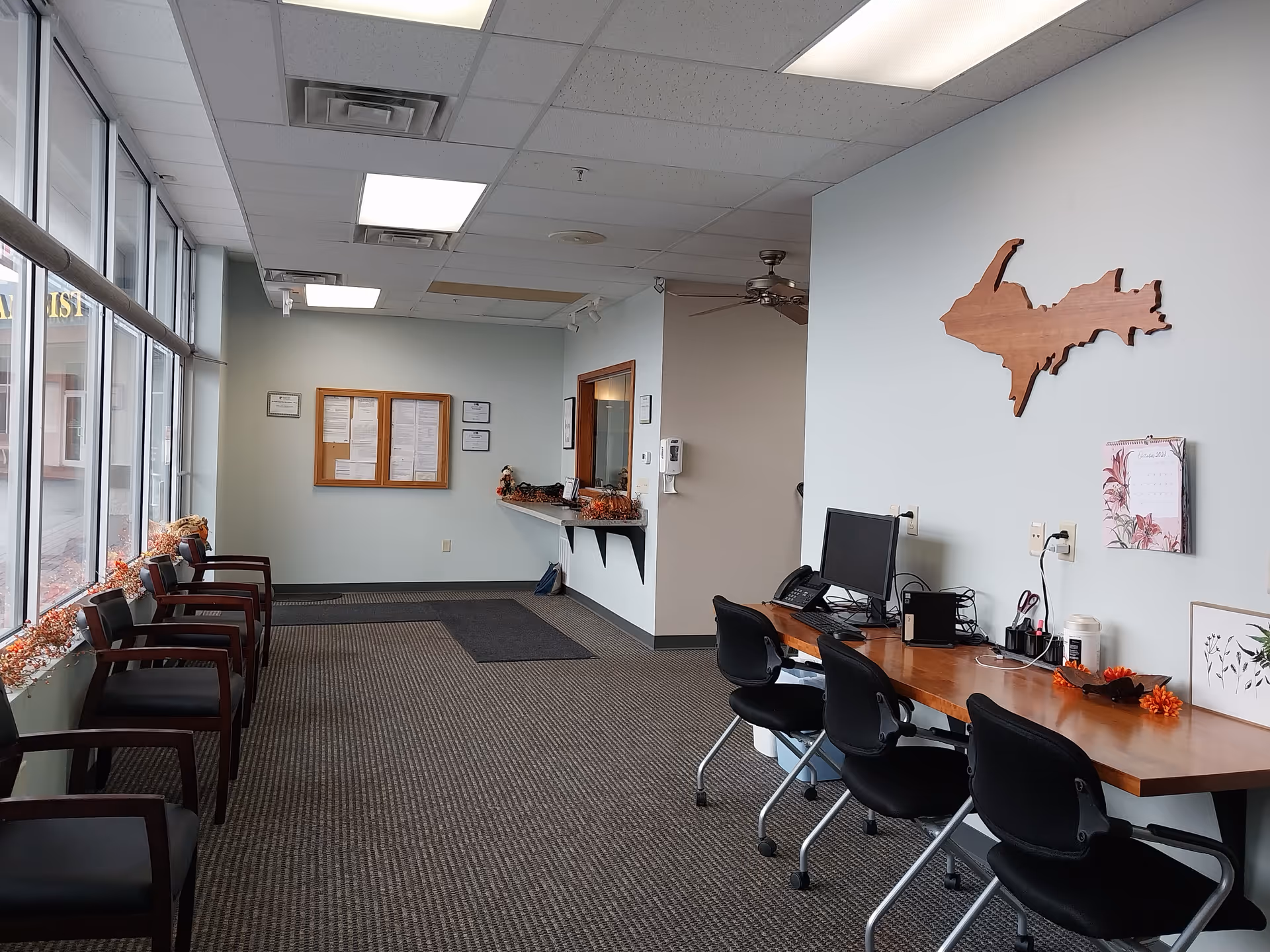 Office reception area with a row of chairs, windows, and a long desk with computers.