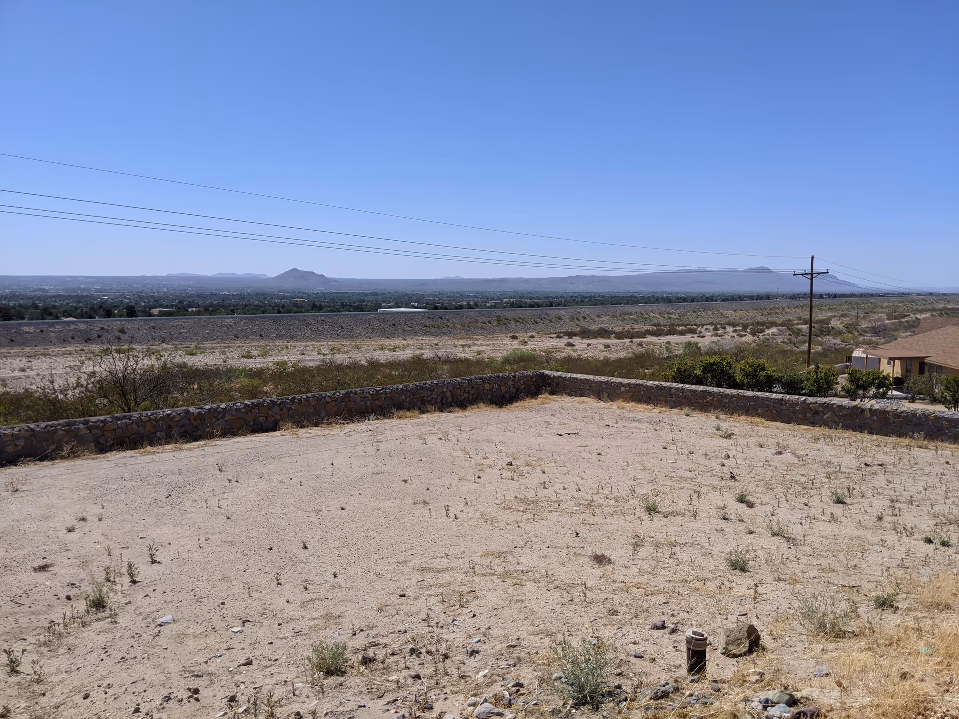 Desert landscape with a low stone wall, power lines, sparse vegetation, and distant houses.