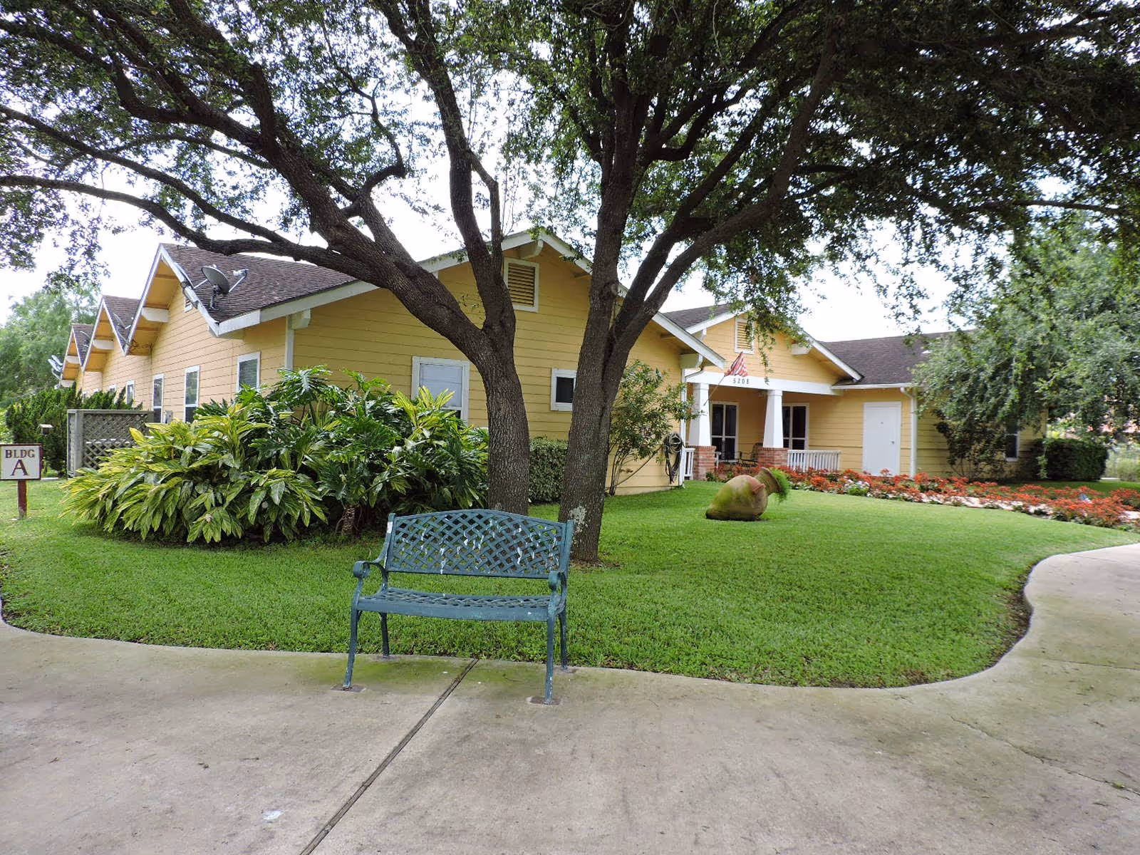 A yellow single-story building with a brown roof surrounded by green grass, trees, and bushes. There is a metal bench in the foreground on a concrete path, and a sign indicating 'BLDG A' near the building. The building has a small porch with white columns and an American flag hanging near the entrance.