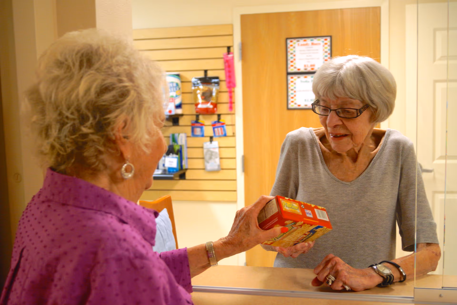 Two elderly women interacting at a counter inside a facility. One woman with short gray hair and glasses is behind the counter, receiving a box of food from another woman with curly gray hair wearing a purple shirt. The background shows a door and a wall with a display rack holding various items.
