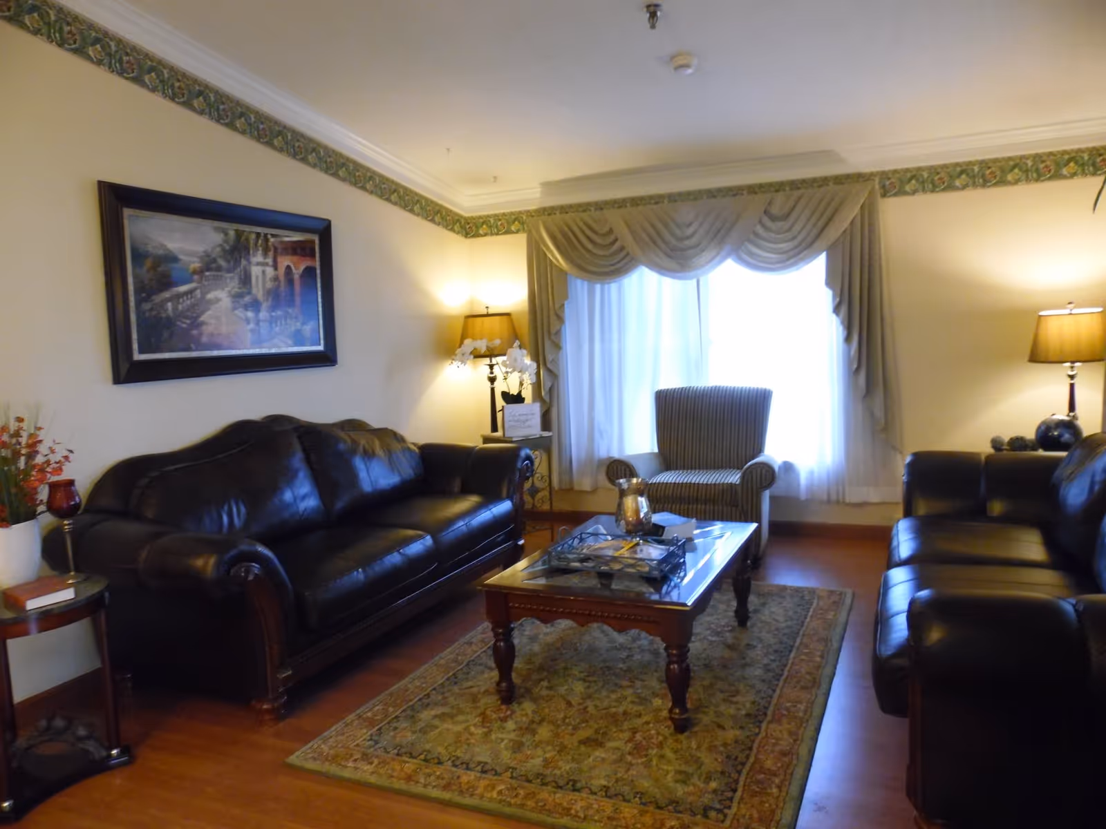 Cozy senior living lounge with leather sofas, a glass coffee table, and draped windows.