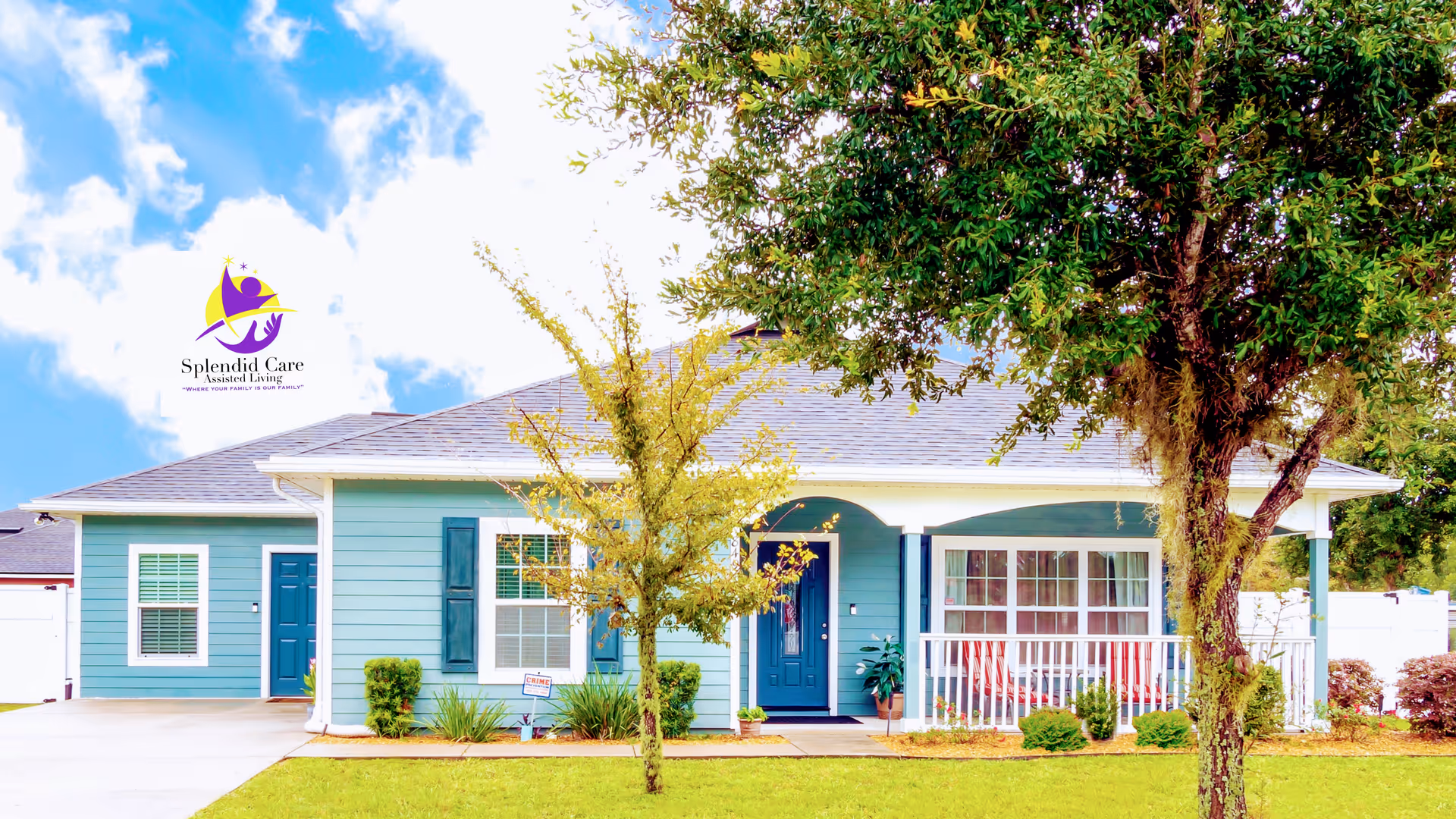 Exterior view of a single-story blue house with white trim and a gray roof, featuring a front porch with white railings, two trees in the front yard, and a clear blue sky with some clouds.