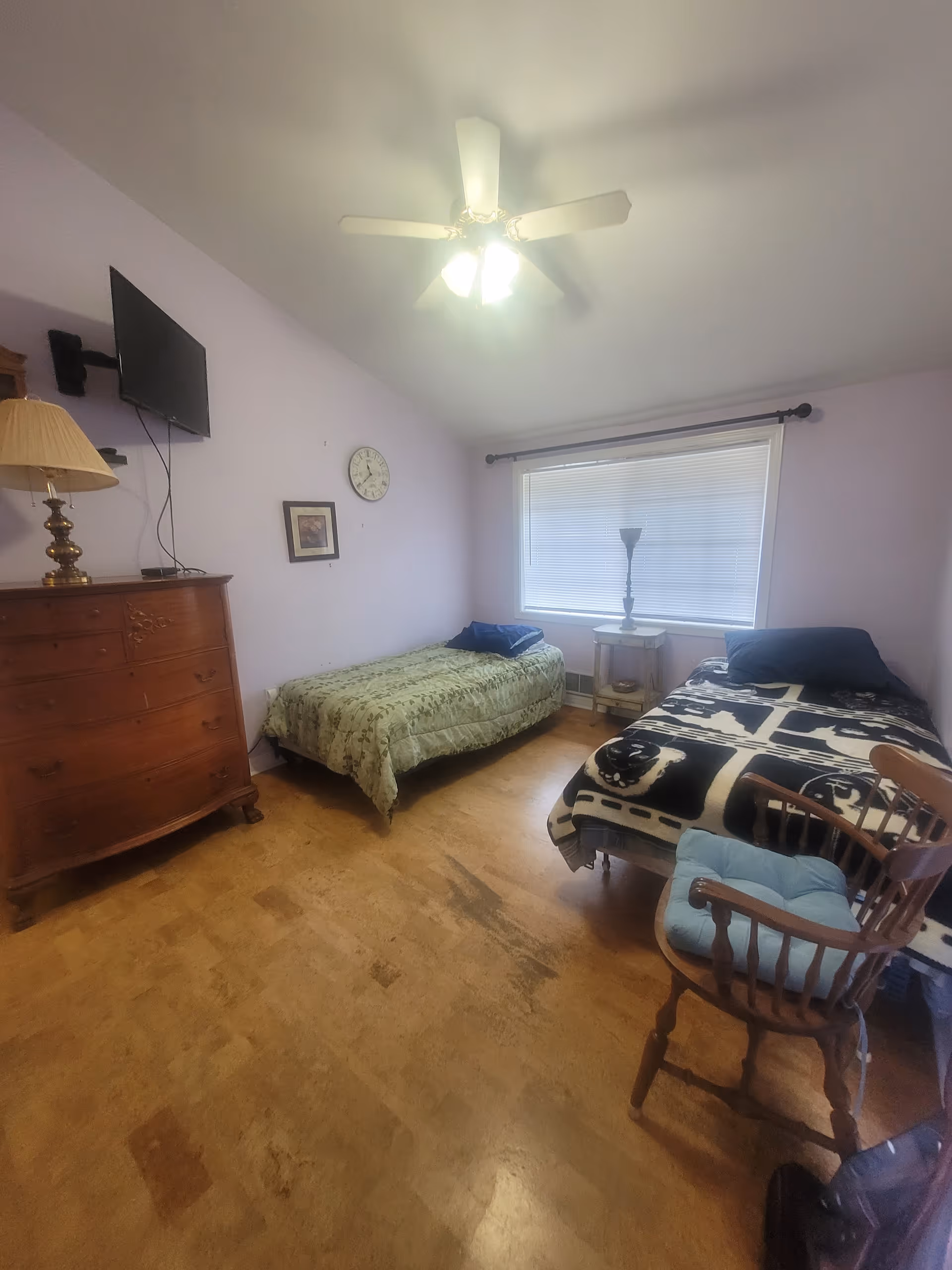 A bedroom with two beds in a senior facility, dresser, window blinds, and ceiling fan.